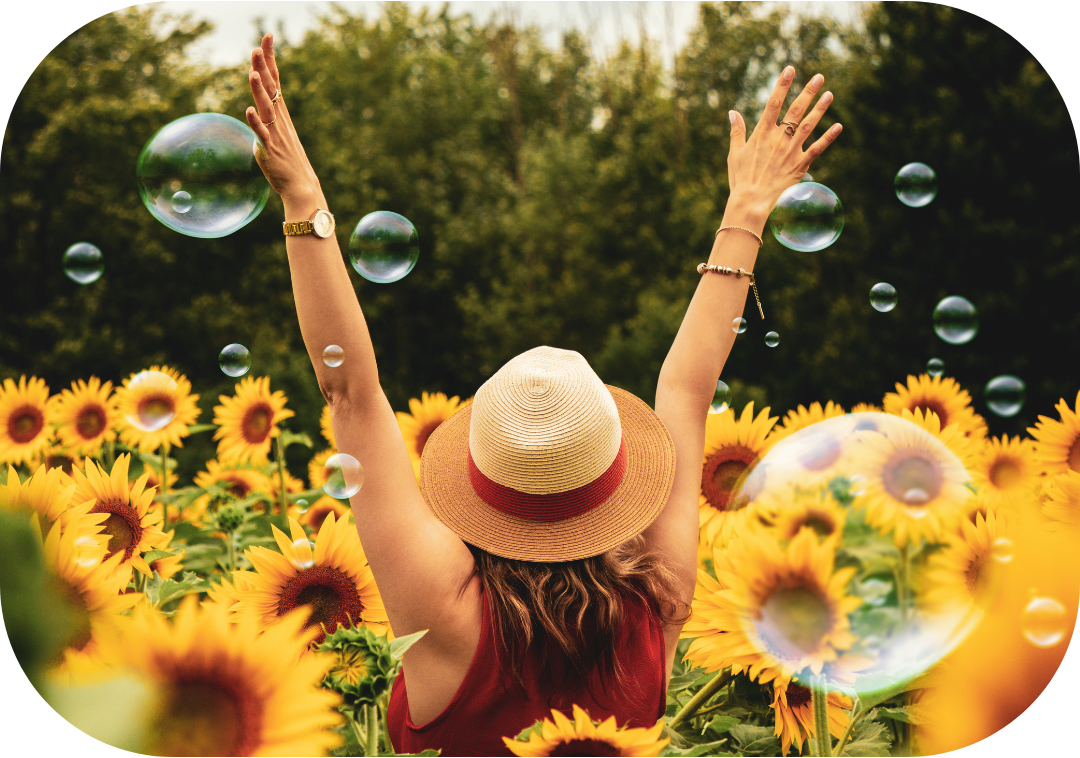 Femme bien dans sa tête et dans son corps qui lèvent les bras au ciel dans un champ de tournesols entourée de bulles de savon