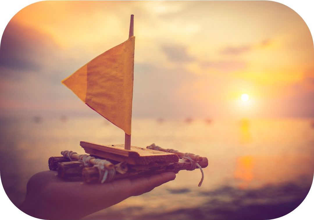 Photo représentant un petit radeau en bois qui tient une main, prêt à être déposé sur l'eau au coucher du soleil.