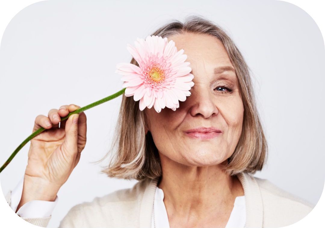 Photo représentant une femme d'environ 60 ans maquillée et avec le sourire qui tient une fleur rose devant sa moitié de visage.